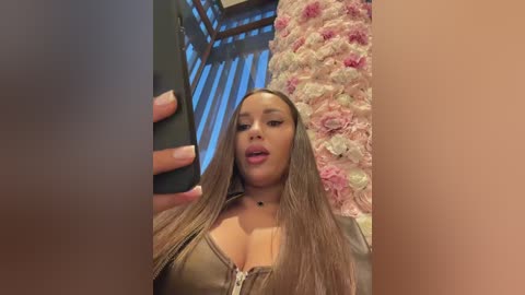 Video of a light-skinned woman with long, straight brown hair, wearing a low-cut, shiny, olive-green top, posing indoors near a floral-patterned wall and a black smartphone.