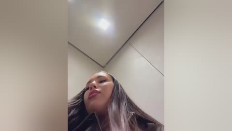 A low-angle video of a woman with long, straight brown hair, fair skin, and full lips, captured from below, looking up. She's wearing a dark jacket. The background is a plain, beige wall with a small light fixture above her head.