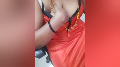 A close-up video of a woman's cleavage in a red satin dress, with black lace, unclasped by a man's hand. She wears a gold necklace and bracelet. Background is blurred.