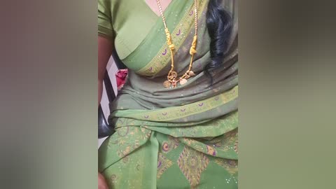 Video of a woman wearing a green sari with intricate yellow embroidery, adorned with gold jewelry, sitting on a black chair with a white and red cushion.