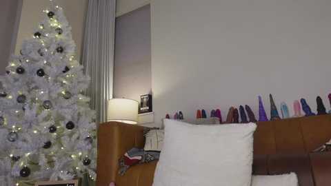 A video of a cozy living room with a lit Christmas tree and a plush sofa adorned with colorful vibrators, a white throw pillow, and a brown blanket.