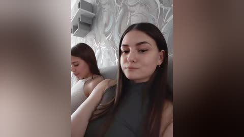 A video of two young women, one with long dark hair, wearing a grey top, sitting in a modern, minimalist interior with grey walls and abstract art. The background is blurred, emphasizing the foreground subjects.