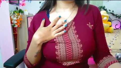 Video of a woman with medium skin tone, wearing a maroon long-sleeve blouse with intricate embroidery, blue nail polish, and a small tattoo on her right wrist. Background includes a pink chair, yellow balloons, and floral arrangements.