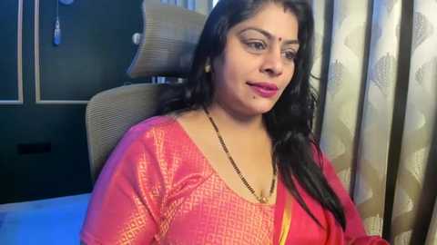 Video of a middle-aged woman with long black hair, wearing a pink saree with gold embroidery, seated on a beige chair, in a dimly lit room with green walls and patterned curtains.