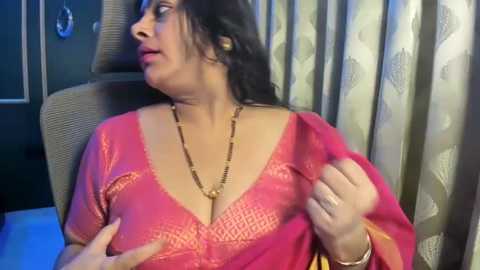 Media: Video of a middle-aged South Asian woman in a bright pink blouse, adjusting her chest area, with a patterned curtain and dark wall in the background.