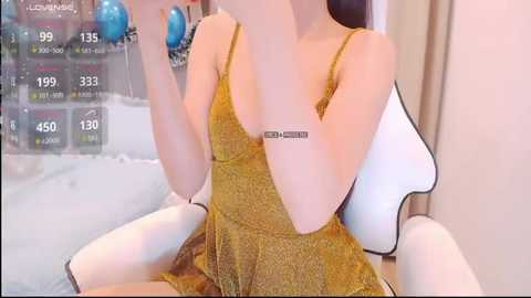 A video of an Asian woman wearing a mustard-yellow sleeveless dress, sitting on a white gaming chair. She's holding a blue ball in her right hand. The background shows a TV displaying financial data.