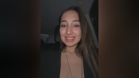 A video of a smiling Latina woman with long brown hair, wearing a black top, captured indoors with dim lighting.