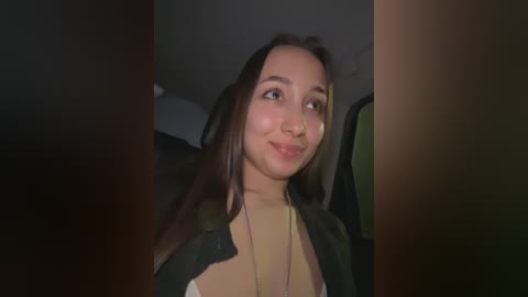 Video of a young woman with light brown skin, long dark hair, wearing a beige top and a black jacket, smiling slightly, sitting in a dimly lit car.