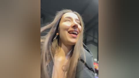 Video of a young woman with long, straight hair, smiling widely, wearing a dark jacket, standing indoors.