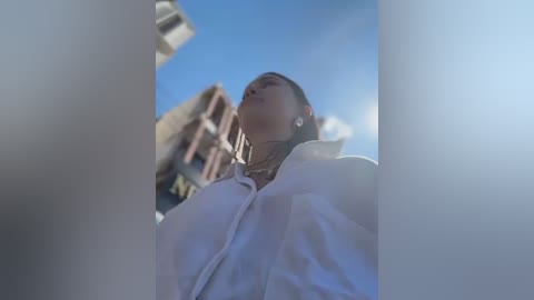 Video of a young woman with dark hair and light skin, wearing a white jacket, looking upwards against a bright blue sky.