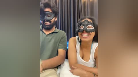 Video of a South Asian couple dressed in black masks, green shirts, and beige pants, seated on a bed with dark curtains in the background.