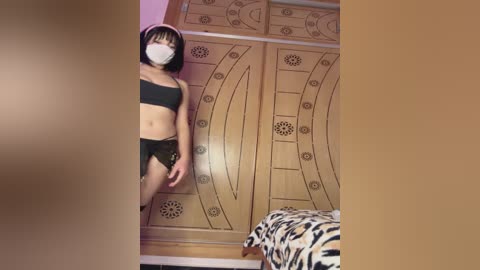 A video of a slender Asian woman in black lingerie and a face mask, standing in a bedroom with a wooden wardrobe and zebra-patterned bedspread.
