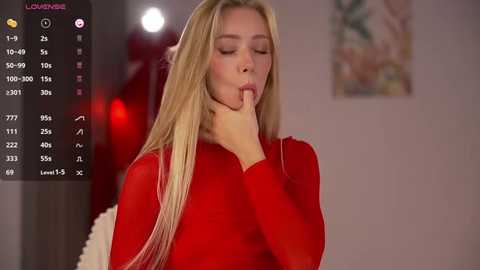 A video of a blonde woman with fair skin, wearing a red top, seductively licking her finger in a dimly-lit room with a blurry, abstract painting on the wall.
