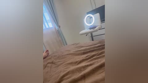 Video of a woman lying face down on a beige bedspread in a dimly lit, minimalistic bedroom with a window covered by sheer curtains. A ring light is mounted on the wall above a white chair, casting a soft glow.