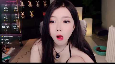 Media: Video of an East Asian woman with long black hair, fair skin, and full lips, wearing a black top, in a dimly-lit room, with a digital overlay of a live stream display.