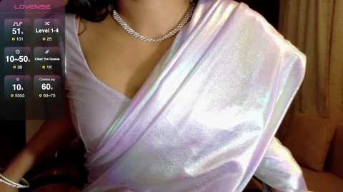 Video of a woman in a shimmering, pastel pink sari, adorned with a diamond necklace, indoors with dim lighting, displaying a social media game overlay.