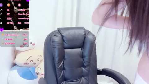 A video of a woman in a white tank top, with long dark hair, standing near a black leather office chair. Background includes a virtual chatroom screen with user avatars and chat text.