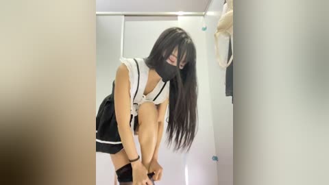 Video of an Asian woman with long black hair, wearing a white blouse and black skirt, bending over in a white-walled, beige-carpeted room, wearing a black face mask.