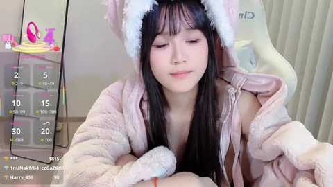A video of a young Asian woman with long black hair, wearing a pink, furry animal hat and robe, lying on a bed, with a digital clock showing 15:20.