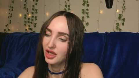 Media: A video of a young woman with long, dark hair and fair skin, wearing a black choker, sitting on a deep blue velvet couch. The background features fairy lights and green ivy, creating a cozy, intimate atmosphere.