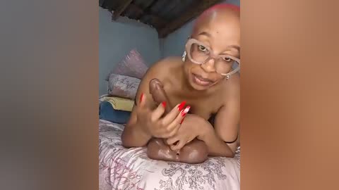 A video of a nude, light-skinned transgender woman with red hair and glasses, holding a large, erect penis. She sits on a bed with floral sheets in a dimly lit, rustic room with a wooden ceiling.