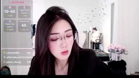 A video of a young East Asian woman with fair skin, long black hair, and clear glasses, wearing a black jacket, seated in a modern, minimalist room with a digital display and floral arrangement in the background.