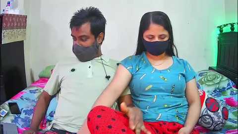 Video of a South Asian couple sitting on a bed with colorful bedding, wearing face masks, the man in a white shirt and the woman in a blue t-shirt.