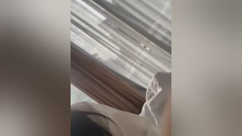 Video of a person in a white shirt with a dark collar, viewed from a low angle. The background features a window with semi-transparent curtains allowing daylight to filter through. The image is slightly blurred, with a soft focus.