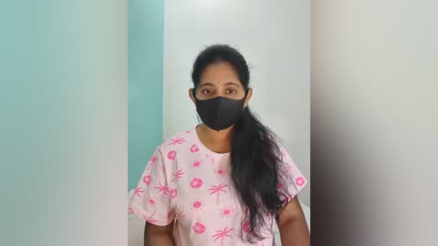 Video of a South Asian woman with medium brown skin and long black hair, wearing a pink floral-patterned hospital gown and a black face mask. The background features a white wall and a teal wall.