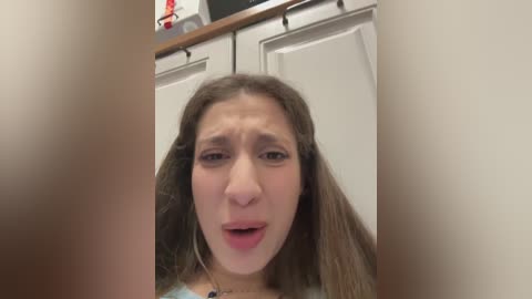 Media: Video of a young woman with light skin and long brown hair, wearing a blue top, looking distressed with open mouth and furrowed brows. Background features white cabinets and a small red object on a countertop.