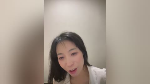 A video of a young Asian woman with straight black hair and light skin, wearing a white shirt, smiling with her mouth slightly open. The background is a plain beige wall.