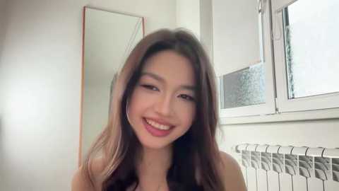 Video of an East Asian woman with long, dark brown hair, smiling, in a bright, white room with a window and radiator.