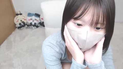 A video of an East Asian woman with shoulder-length black hair, wearing a light gray sweater and a face mask, sitting on a white couch in a minimalist room. In the background, stuffed animals are visible.