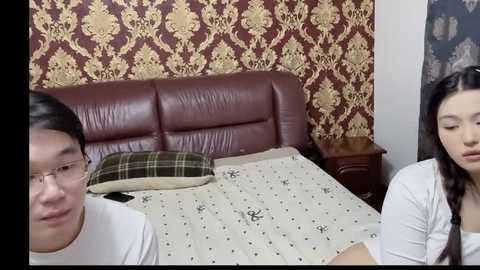 Media: A video showing a young Asian couple in a bedroom with a dark brown leather headboard, red and gold damask wallpaper, and a white bed with a green plaid pillow.