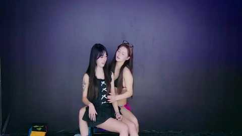 Video of two young East Asian women with long hair, dressed in revealing black corsets and pink panties, sitting intimately on a dark floor.