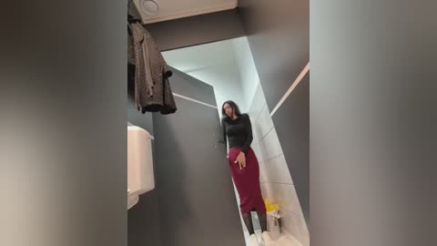 Video of a woman in a restroom, wearing a black long-sleeve top and a red skirt, holding a black jacket. Gray walls, white ceiling, and a white sink in the background.