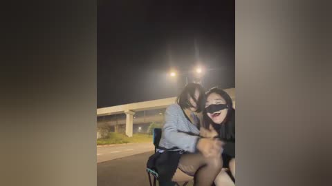 Video of a nighttime scene featuring two Asian women, one riding on a man's shoulders, smiling with a black blindfold. They are on a dark road beside a highway, with bright streetlights illuminating them.