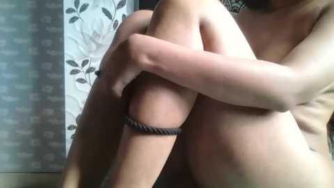 A video of a nude person with light skin, hugging their knees, wearing a grey rope around their left ankle. Background features a floral-patterned wall and a blurred white and grey textured wall.
