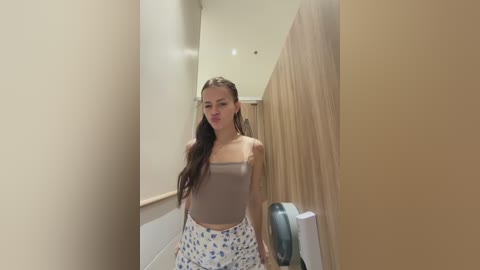 Video of a young woman with long brown hair, wearing a brown tank top and blue-patterned pants, standing in a narrow, beige-walled hallway with a wooden door and hand sanitizer dispenser.