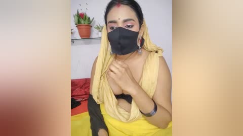 Video of a South Asian woman with medium skin tone, wearing a yellow saree, black face mask, and red bindi, holding her chest, in a simple indoor setting with potted plants.