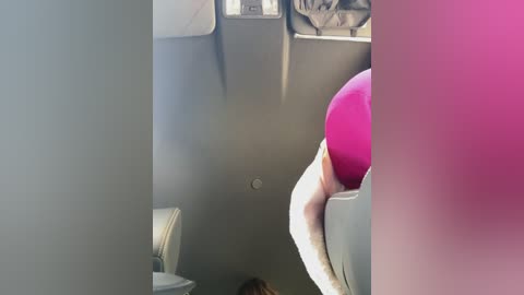 A video of a woman in a bright pink top, leaning over a gray airplane seat, with a blurred view of the seat and window.