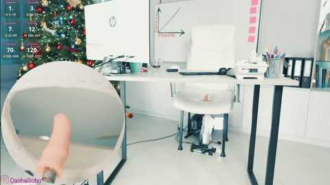 Video of a modern, minimalist office with a white desk, white chair, and a large whiteboard. A hand holds a VR headset in the foreground. A decorated Christmas tree stands in the background.