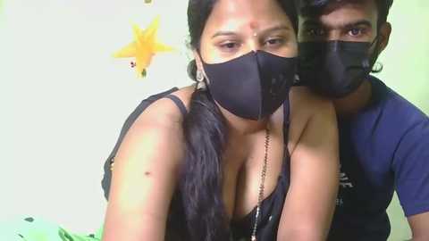 Video of a South Asian woman with medium skin tone, long black hair in braids, wearing a black mask and revealing black bra, and a man behind her, both in a room with yellow star decorations on a light green wall.