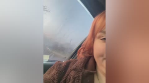 Video of a person with short, fiery red hair and light skin, wearing a brown, fuzzy coat, smiling slightly, partially visible through a foggy car window.