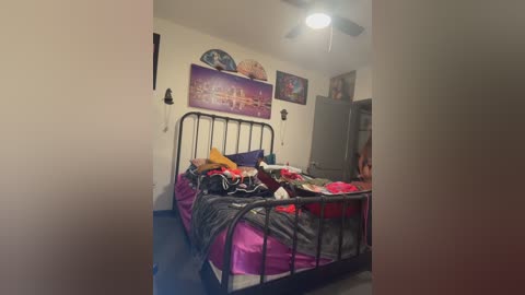 Video of a messy bedroom with a black metal bed, pink and purple sheets, cluttered with clothes, framed photos, and a large refrigerator on the right.