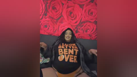 Video of a plus-size woman with dark hair, fair skin, and a big smile, wearing a black hoodie with \"LOOK BETTER BE OVER\" and orange panties, against a red rose background.