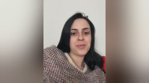 Video of a young woman with long black hair, fair skin, and a subtle smile, wearing a textured, multicolored knit sweater against a plain beige background.