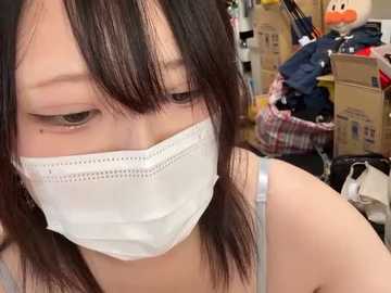 Media: Video of an Asian woman with long black hair, wearing a white surgical mask, a gray tank top, and sitting in a cluttered room filled with cardboard boxes and clothes.
