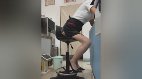 Video of an Asian woman in a white shirt and black skirt, leaning against a blue wall in a small, cluttered office. Black stiletto heels, visible panties, computer monitor, and a trash can are present.