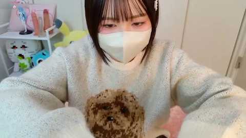 Video of an Asian woman with short black hair, wearing a white face mask and beige sweater, holding a small, brown, curly-haired dog. Background includes a white wall, a pink storage bin, and plush toys.